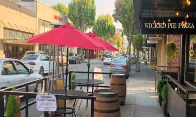 Outdoor café seating with a pink umbrella on a sidewalk beside barrels and a Wicked Pie Pizza storefront on the right.