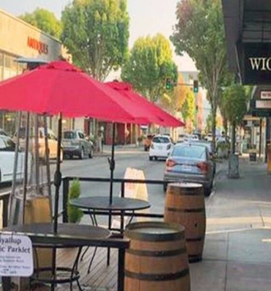Outdoor café seating with a pink umbrella on a sidewalk beside barrels and a Wicked Pie Pizza storefront on the right.