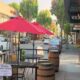 Outdoor café seating with a pink umbrella on a sidewalk beside barrels and a Wicked Pie Pizza storefront on the right.