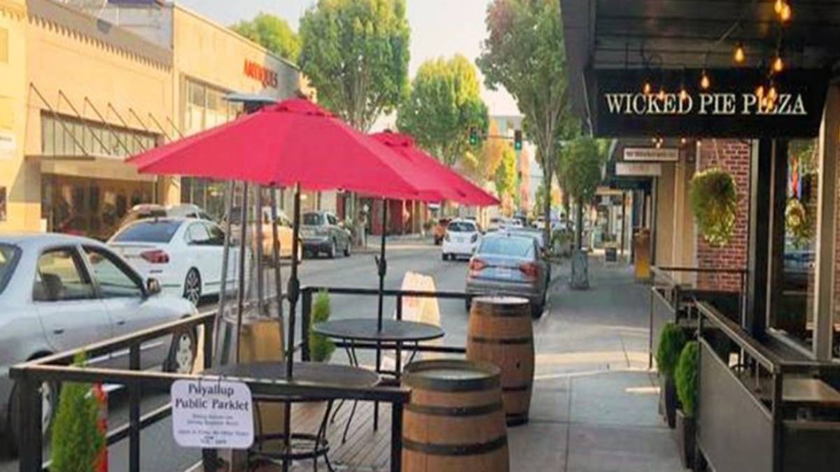 Outdoor café seating with a pink umbrella on a sidewalk beside barrels and a Wicked Pie Pizza storefront on the right.