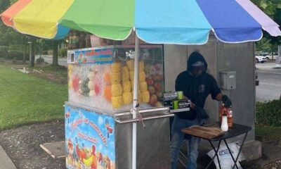 Fruit juice stand with a rainbow umbrella; vendor in a dark hoodie prepares drinks beside a fruit display on ice.