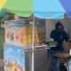 Fruit juice stand with a rainbow umbrella; vendor in a dark hoodie prepares drinks beside a fruit display on ice.