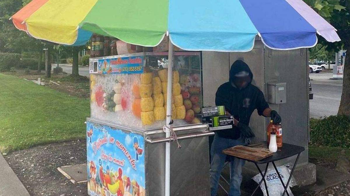 Fruit juice stand with a rainbow umbrella; vendor in a dark hoodie prepares drinks beside a fruit display on ice.