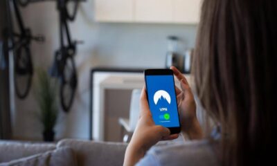 Person sits on a sofa holding a smartphone that shows a VPN app with a blue screen and mountain logo, indicating a connection.