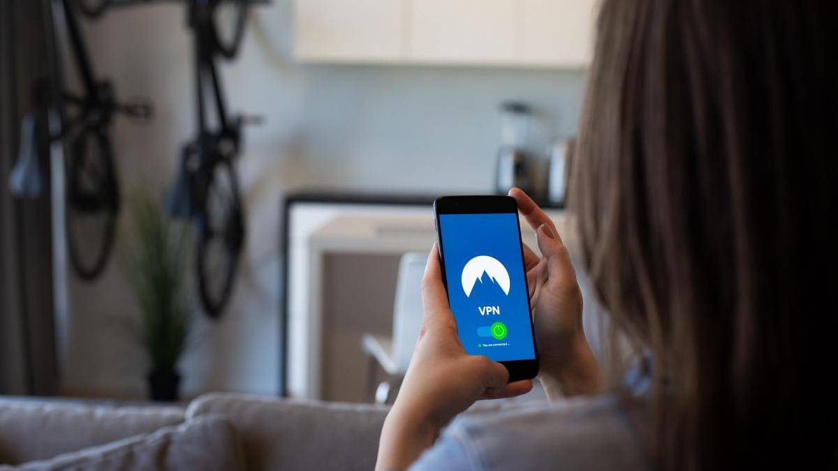 Person sits on a sofa holding a smartphone that shows a VPN app with a blue screen and mountain logo, indicating a connection.