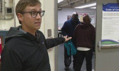 Man with glasses in a black jacket stands at a doorway, gesturing toward a group entering a room with teal bags in hand.