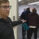 Man with glasses in a black jacket stands at a doorway, gesturing toward a group entering a room with teal bags in hand.