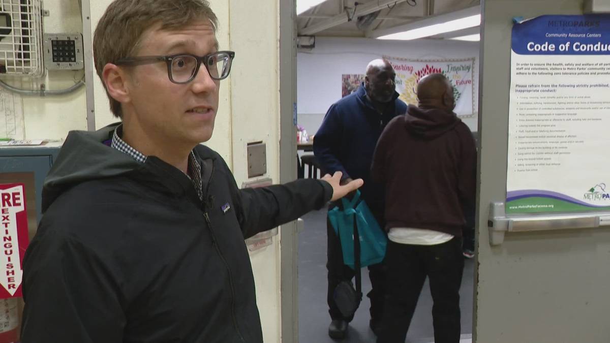 Man with glasses in a black jacket stands at a doorway, gesturing toward a group entering a room with teal bags in hand.