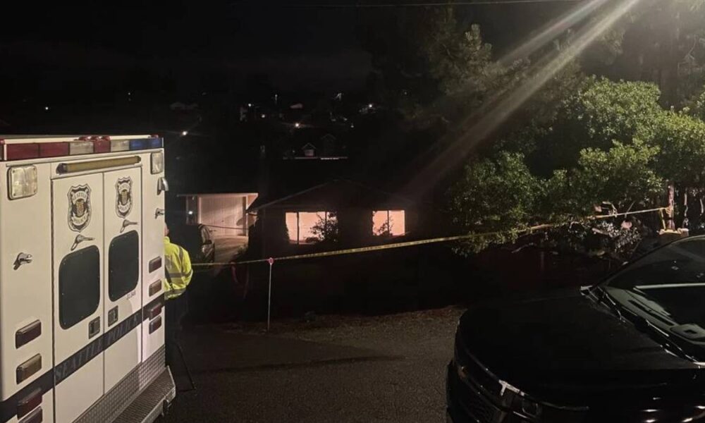 Nighttime emergency scene with an ambulance on the left, a responder in a reflective vest, and yellow caution tape cordoning off a residential area.