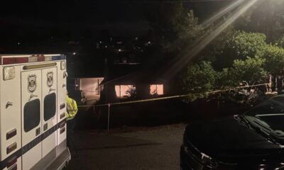 Nighttime emergency scene with an ambulance on the left, a responder in a reflective vest, and yellow caution tape cordoning off a residential area.