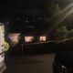 Nighttime emergency scene with an ambulance on the left, a responder in a reflective vest, and yellow caution tape cordoning off a residential area.