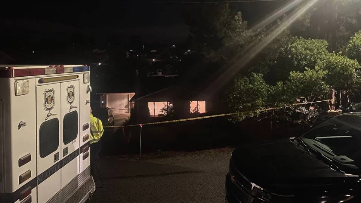 Nighttime emergency scene with an ambulance on the left, a responder in a reflective vest, and yellow caution tape cordoning off a residential area.