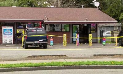 A blue SUV parked in front of a small shop, yellow caution tape stretched across the scene, storefront windows and vending machines visible behind it.