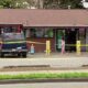 A blue SUV parked in front of a small shop, yellow caution tape stretched across the scene, storefront windows and vending machines visible behind it.