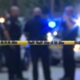 Yellow crime scene tape stretches across the foreground while blurred police officers and flashing blue lights appear in the background.