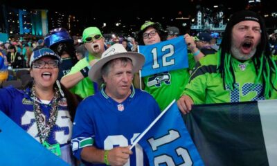 Crowd of Seattle Seahawks fans cheering at a stadium, wearing blue and green, waving flags and wearing jersey number 12.