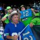 Crowd of Seattle Seahawks fans cheering at a stadium, wearing blue and green, waving flags and wearing jersey number 12.