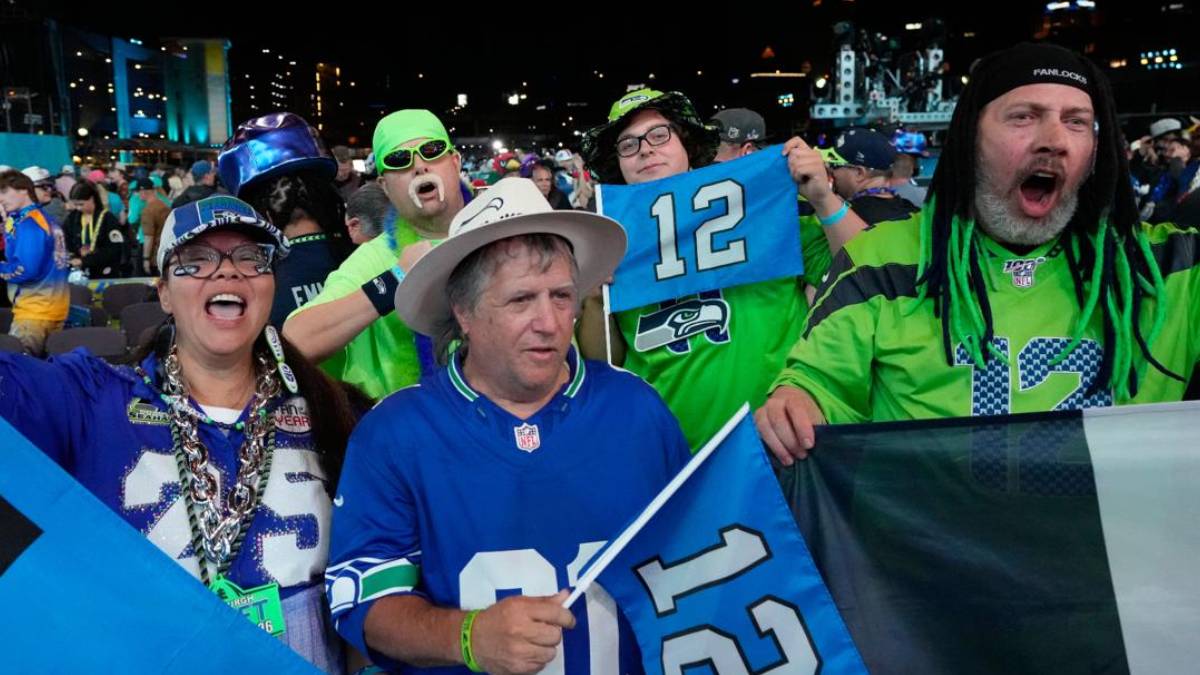 Crowd of Seattle Seahawks fans cheering at a stadium, wearing blue and green, waving flags and wearing jersey number 12.