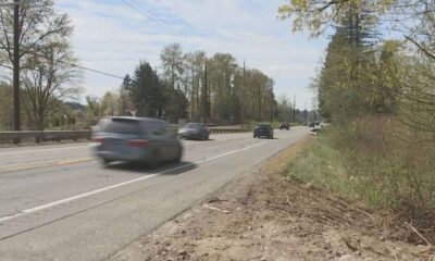 Blurred cars speeding down a rural, two-lane road with a guardrail and trees lining the roadside under a blue sky.