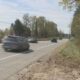 Blurred cars speeding down a rural, two-lane road with a guardrail and trees lining the roadside under a blue sky.