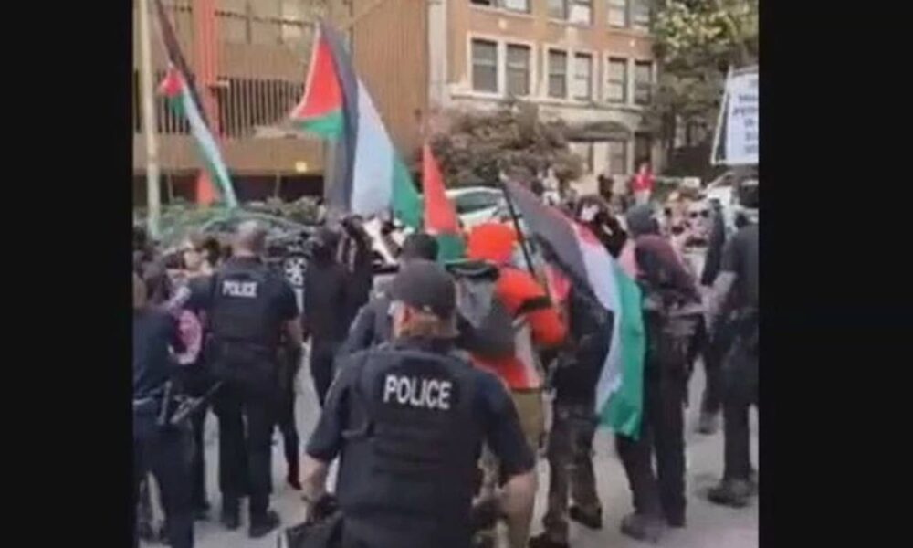 Protest on a city street with police in tactical vests confronting demonstrators, many waving Palestinian flags and banners in the background.