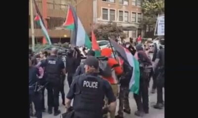 Protest on a city street with police in tactical vests confronting demonstrators, many waving Palestinian flags and banners in the background.
