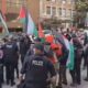 Protest on a city street with police in tactical vests confronting demonstrators, many waving Palestinian flags and banners in the background.