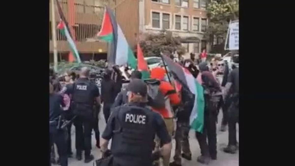 Protest on a city street with police in tactical vests confronting demonstrators, many waving Palestinian flags and banners in the background.
