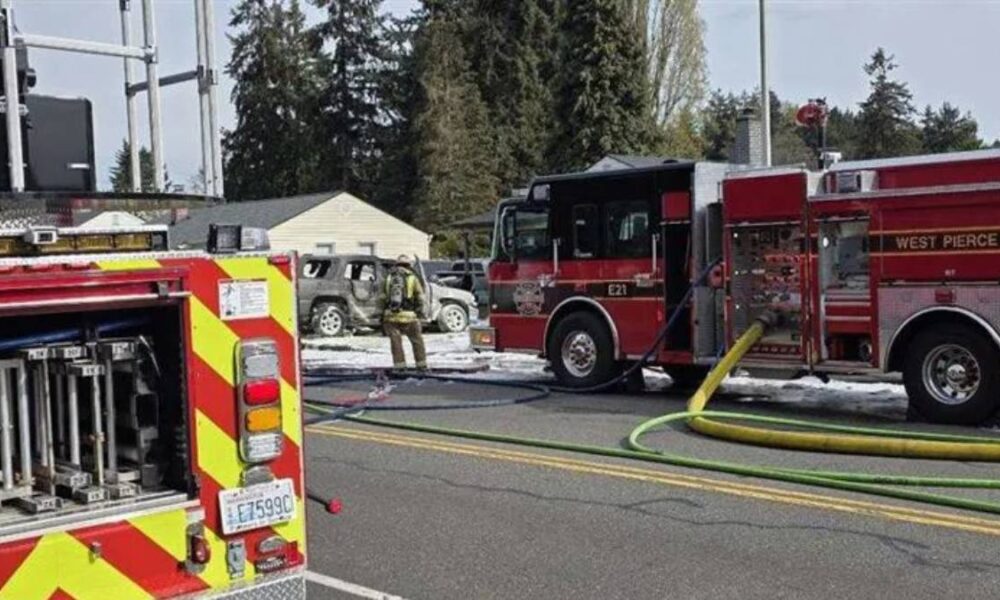 Fire trucks on a street with hoses laid out, firefighters attending a scene near a small damaged vehicle in the background.