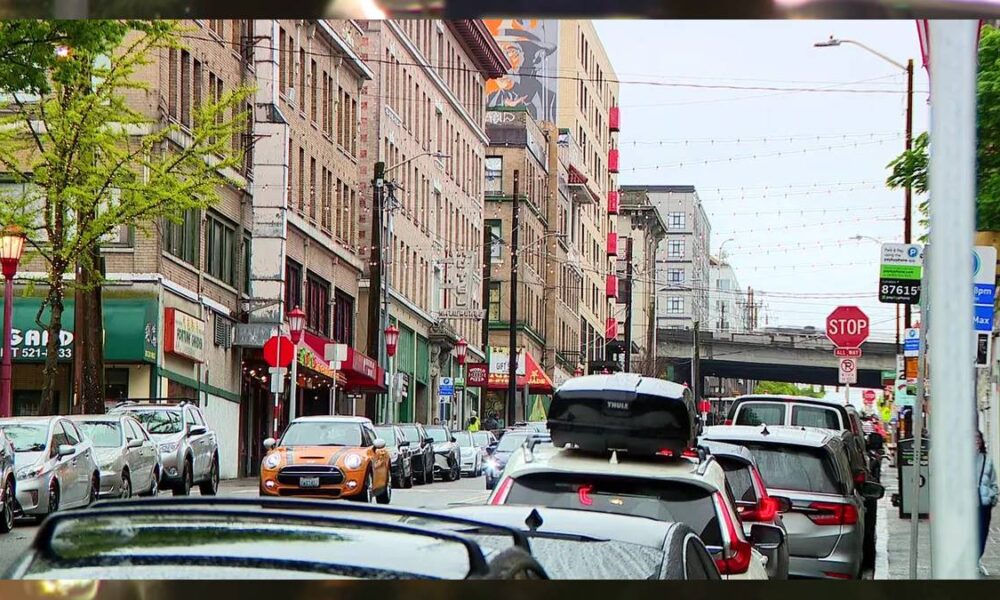 Busy urban street with parked cars along storefronts and a brick building row; an orange Mini Cooper drives toward the camera under string lights.