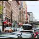 Busy urban street with parked cars along storefronts and a brick building row; an orange Mini Cooper drives toward the camera under string lights.