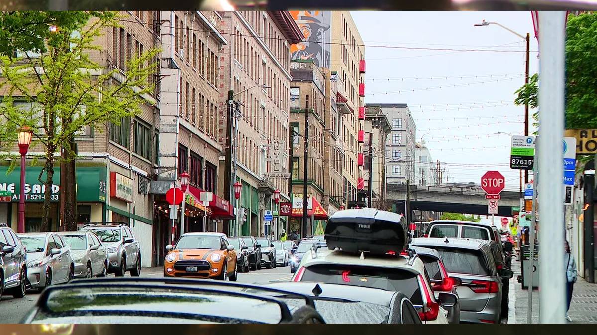 Busy urban street with parked cars along storefronts and a brick building row; an orange Mini Cooper drives toward the camera under string lights.