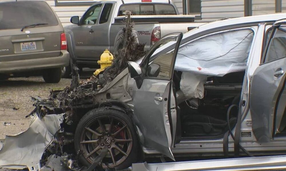 Damaged silver car with extensive front-end destruction and deployed airbags after a crash; open doors and bent front wheel visible, parked beside other vehicles on a street lot.