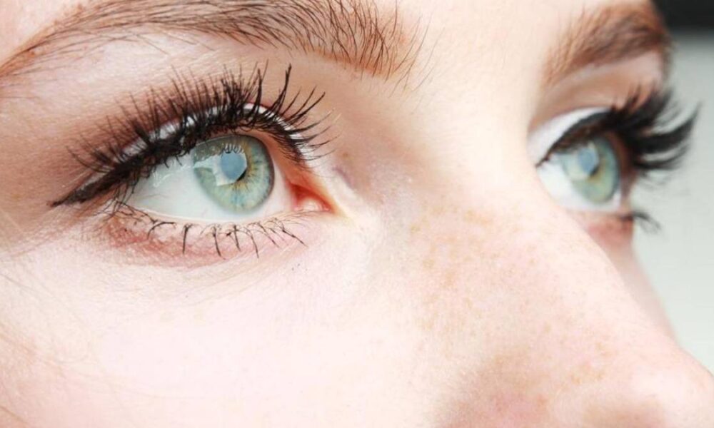 Close-up of a person's blue-green eyes with long black eyelashes and light freckles on the nose and cheek.
