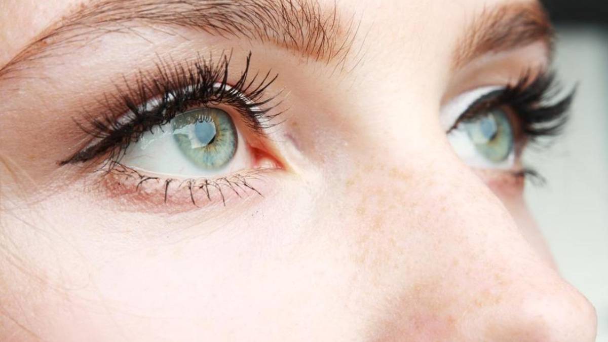 Close-up of a person's blue-green eyes with long black eyelashes and light freckles on the nose and cheek.