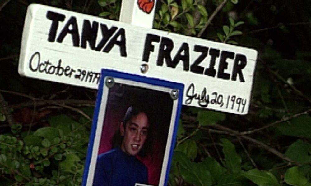 Roadside memorial sign with a hanging portrait photo of a young woman among leafy bushes.