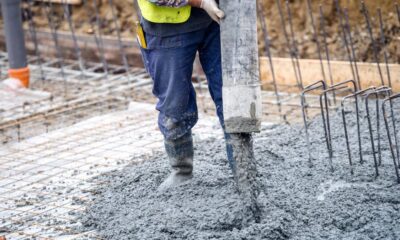 Construction worker in blue pants and yellow vest pours wet concrete over a rebar grid at a building site.