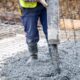 Construction worker in blue pants and yellow vest pours wet concrete over a rebar grid at a building site.