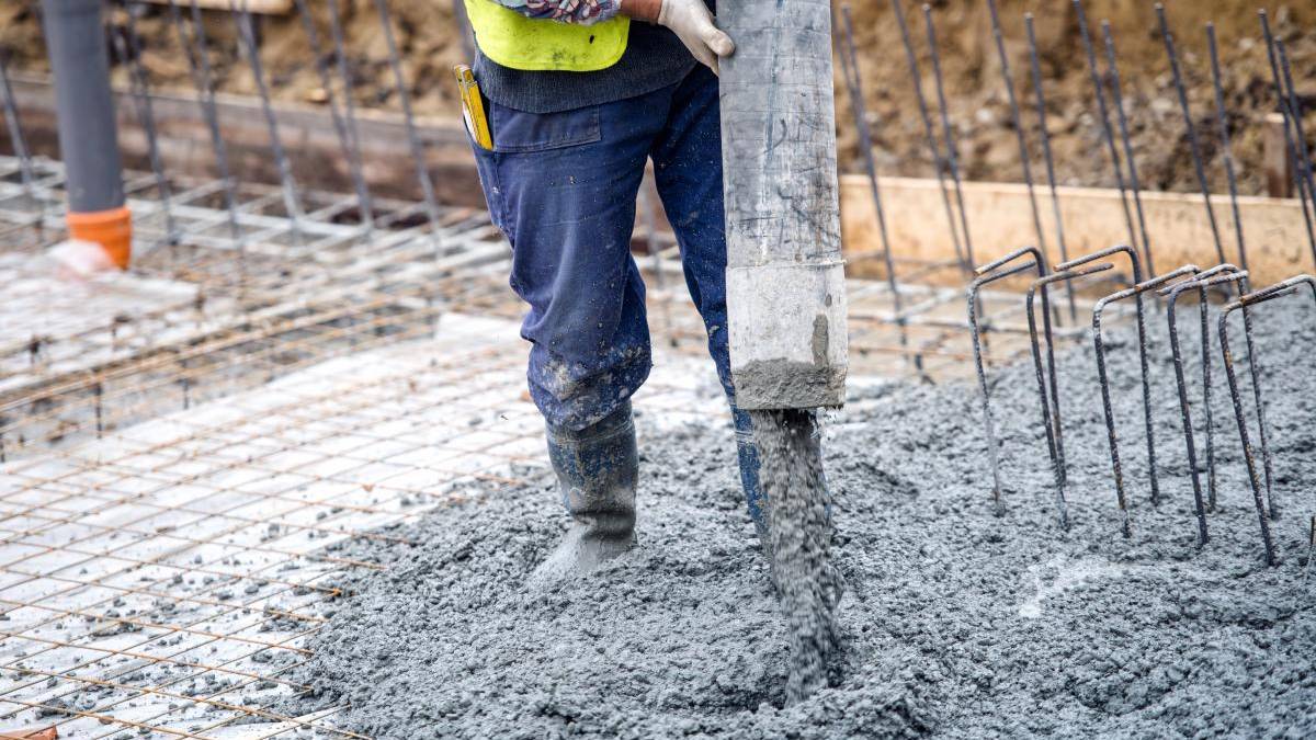 Construction worker in blue pants and yellow vest pours wet concrete over a rebar grid at a building site.