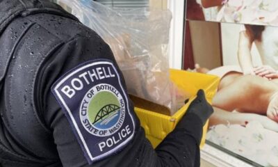 Close-up of a Bothell Police officer in a dark jacket holding a yellow plastic bin near a storefront window with a posted image.