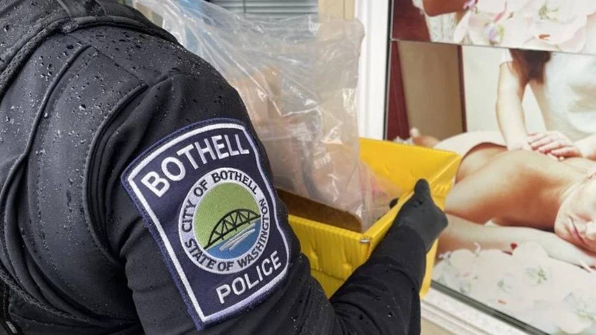 Close-up of a Bothell Police officer in a dark jacket holding a yellow plastic bin near a storefront window with a posted image.