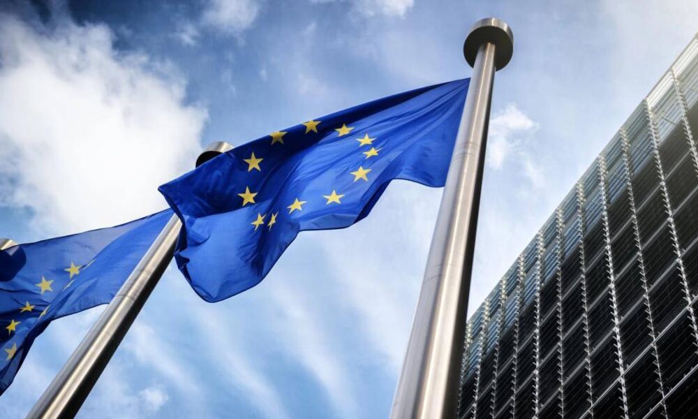 European Union flag waving on a tall pole beside a modern glass office building under a blue sky