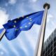 European Union flag waving on a tall pole beside a modern glass office building under a blue sky