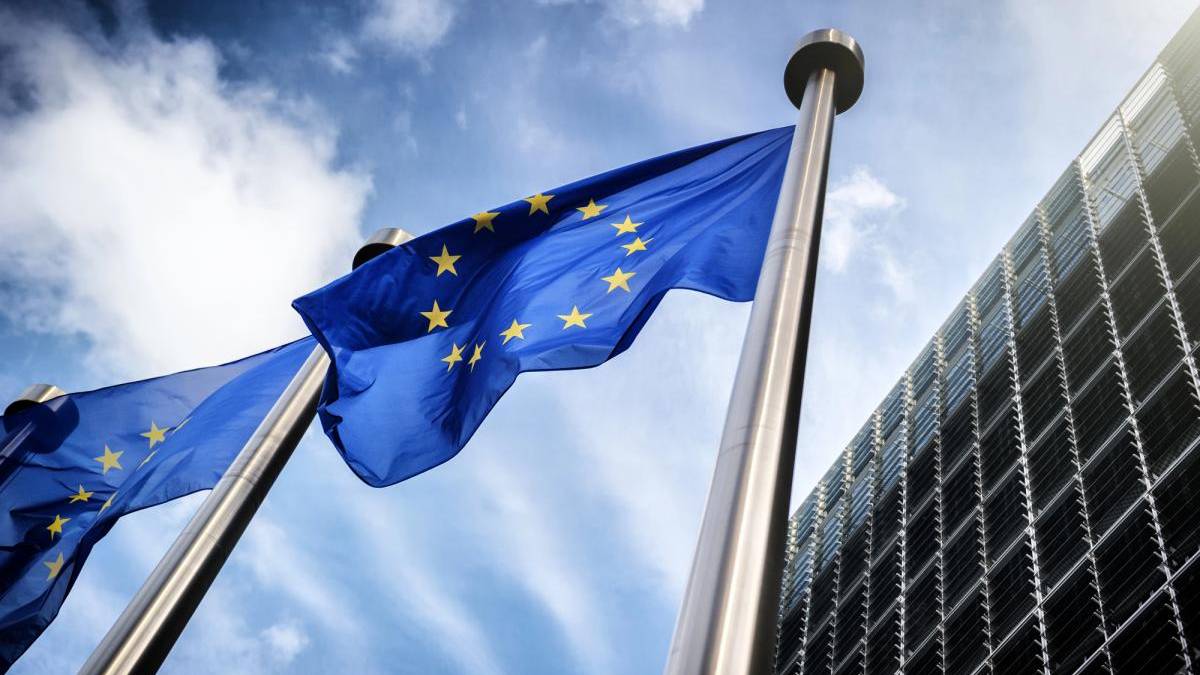 European Union flag waving on a tall pole beside a modern glass office building under a blue sky