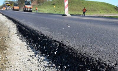 New asphalt being laid on a road under construction with workers and machinery in the background, in a rural setting.