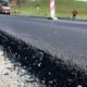 New asphalt being laid on a road under construction with workers and machinery in the background, in a rural setting.