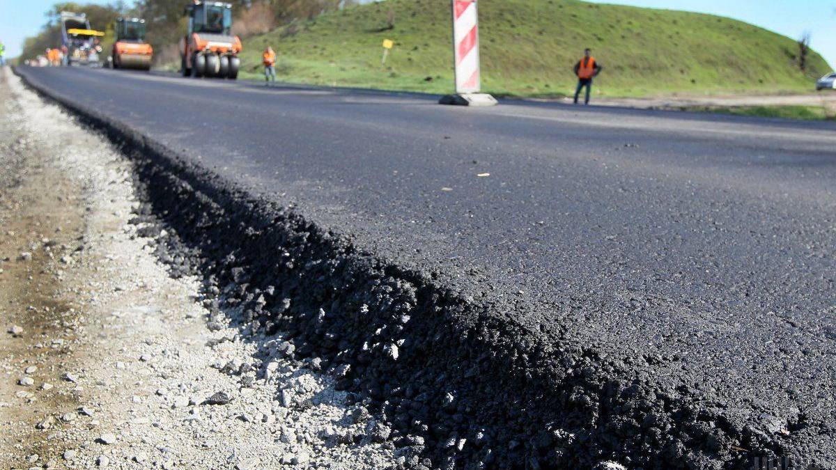 New asphalt being laid on a road under construction with workers and machinery in the background, in a rural setting.