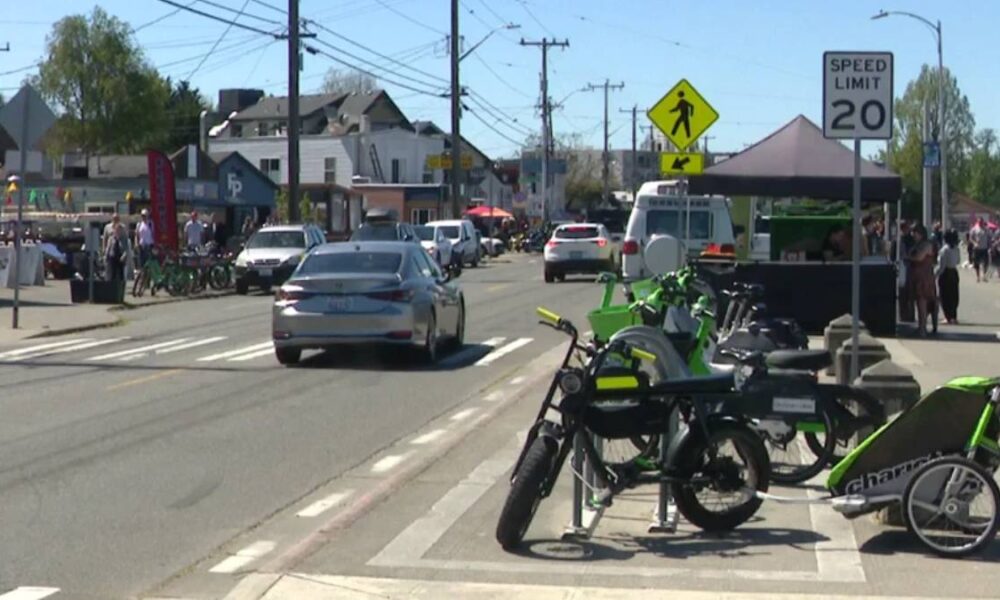 Street scene with bright green bicycles parked at a curb, market stalls and pedestrians along a sunny downtown road as cars drive by.