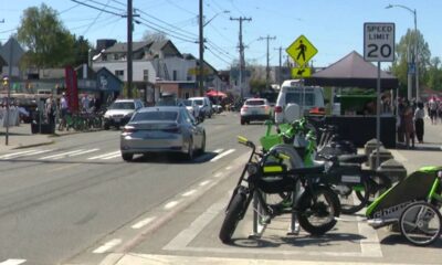 Street scene with bright green bicycles parked at a curb, market stalls and pedestrians along a sunny downtown road as cars drive by.