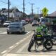 Street scene with bright green bicycles parked at a curb, market stalls and pedestrians along a sunny downtown road as cars drive by.